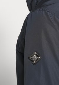 Mörkblå jacka med en slät textur, diamantformad logotypflik med metalldetaljer på vänster axel. Minimalistisk design och rena linjer.