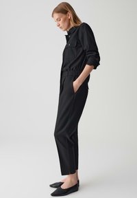 Completo due pezzi nero composto da una giacca con colletto e tasca, con vita elastica, abbinata a pantaloni sartoriali e scarpe slip-on nere.