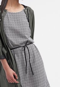 Svartvit geometrisk mönstrad klänning med lös passform, åtdragen i midjan med ett svart band. Olivgrön jacka bärs över den.