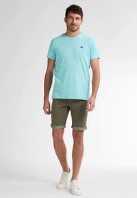 Světlomodré tričko s krátkým rukávem a malým logem na hrudi, v kombinaci se zelenými olivovými šortkami s ohrnutými nohavicemi a bílými teniskami.