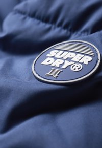 Marine stof met een soepele textuur, voorzien van een ronde, gestikte patch met het label "SUPER DRY" in het wit, met horizontale lijnen en extra tekens.