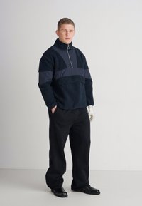 Marineblauwe fleece pullover met hoge kraag, ritssluiting en contrasterende donkere panels; gecombineerd met zwarte broeken en handschoenen.
