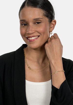 Gold chain necklace with layered design and a rectangular accent. Model wearing matching gold bracelet and hoop earrings. Black blazer, white top.