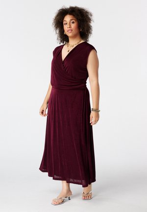 Femme aux cheveux bouclés portant une robe portefeuille bordeaux sans manches et des sandales argentées à talons, accessoirisée de bijoux en or.