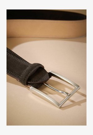 Bruine suède riem met een zilveren rechthoekige gesp, voorzien van gestikte randen en een gladde textuur, ligt op een beige achtergrond.