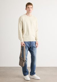 Lys beige genser med ribbede mansjetter, kombinert med blå denimjeans og hvite joggesko. En liten brun crossover-veske holdes i den ene hånden.