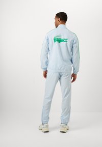 Jachetă sport de culoare albastru deschis, cu un logo verde de crocodil pe spate, asociată cu pantaloni asortați. Materialul are o textură netedă; manșete elastice.
