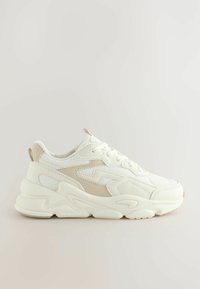 Baskets de sport blanches et beiges en maille et matériaux synthétiques, avec une semelle épaisse, un bout arrondi et des accents texturés.