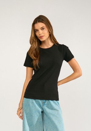 Frau mit langen braunen Haaren, die ein eng anliegendes schwarzes kurzärmeliges Shirt und lockere hellblaue Jeans trägt, posiert mit einer Hand auf der Hüfte.