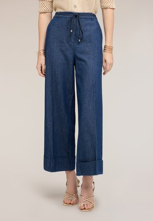 Femme portant un pantalon en denim bleu taille haute à jambes larges avec ourlets retroussés et taille à cordon, associé à des sandales dorées à lanières et des bracelets.