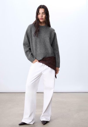Grauer Strickpullover mit rundem Ausschnitt, kombiniert mit weißen weiten Hosen und einem braunen gemusterten Taillengürtel. Spitz zulaufende schwarze Schuhe ergänzen das Outfit.