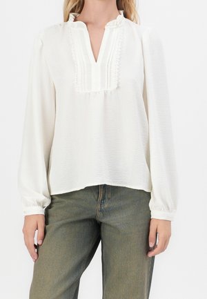 Vrouw draagt een witte blouse met lange mouwen met geplooide, met kant afgewerkte V-hals en olijfgroene high-waist jeans.