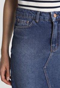 Spijkerrok in een medium blauwe wassing met vijf zakken, hoge taille en contrasterende stiksels. Voorzien van een knoopsluiting en riemlussen.