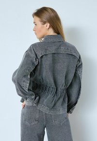 Hallideg halli denimhjakk, millel on pitsitatud vöökoht, nööbitav esiosa ja pikad varrukad. Omab teravat kaelust ja lihtsaid õmblusdetailide.