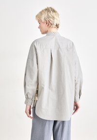 Camicia a righe con bottoni in grigio chiaro e beige. Presenta una vestibilità ampia, spalle cadenti e una fenditura sul retro con dettaglio a bottone. Gli accessori includono dei lacci ai lati.