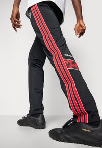 Adidas black athletic pants with red and white side stripes, featuring a geometric pattern and breathable fabric. Black sport shoes complement the look.