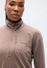 Πουλόβερ από φλις σε ανοιχτό καφέ χρώμα με ψηλό γιακά, μισό φερμουάρ, ριμπ υφή και λεπτομέρεια λογότυπου στο στήθος. Μαλακό φινίρισμα υφάσματος.