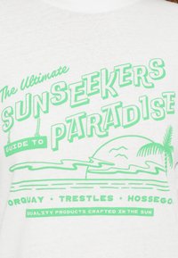 Λευκό μπλουζάκι από ύφασμα με πράσινο κείμενο. Ο σχεδιασμός περιλαμβάνει το "Ο απόλυτος οδηγός των λάτρεων του ήλιου προς τον �παράδεισο" με γραφικά παραλίας και φοίνικες.