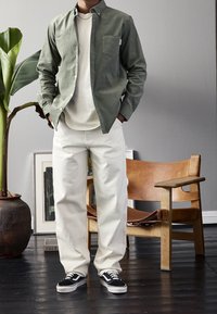 Camisa de botones verde oliva, camiseta blanca, pantalones blanco roto, zapatillas negro y blanco. Silla de madera y planta verde al fondo.