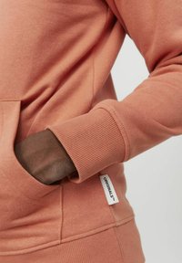 Sudadera rosa clara con puños acanalados y un bolsillo tipo canguro. Presenta una pequeña etiqueta con la inscripción "ORIGINALS" en la costura lateral. Textura suave.