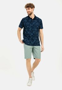 Marineblaues Poloshirt mit einem Blattmuster, kombiniert mit hellgrünen Shorts. Das Outfit wird mit weißen Sneakers abgerundet.