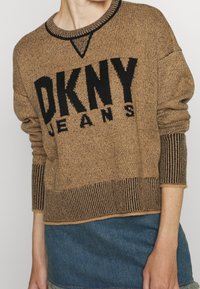 Pull en maille marron avec le logo noir "DKNY JEANS". Caractéristiques : col et poignets côtelés, design texturé et légèrement surdimensionné.