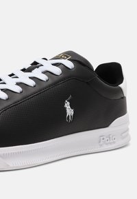 Baskets noires avec lacets blancs, semelle blanche embossée de ""Polo,"" et un logo blanc de joueur de polo sur le côté.