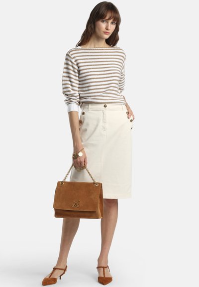 Femme debout portant un pull à rayures marron et blanc, une jupe crème boutonnée, un sac à main en daim marron et des talons marron assortis.