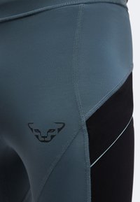Gros plan sur des leggings de sport gris et noirs avec un petit logo stylisé de visage de chat noir sur la cuisse.