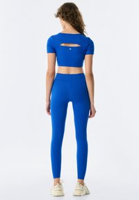 Top blu cropped con maniche corte e dettagli a taglio sulla schiena, abbinato a leggings blu a vita alta. Tessuto liscio ed elastico.