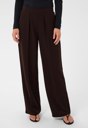 Personne portant un pantalon large marron foncé taille haute avec des plis devant, associé à un haut noir à manches longues et des sandales noires à brides entre les orteils.