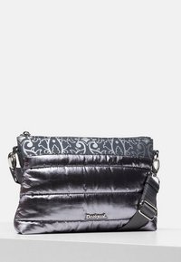 Wypukła metaliczna srebrna torba z szarym wzorzystym materiałowym wierzchem, regulowanym paskiem i zamkiem błyskawicznym. Na przodzie znajduje się logo "Desigual".