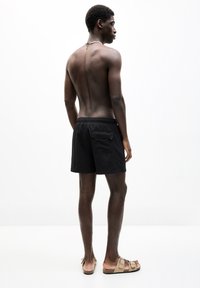 Schwarze Badeshorts mit einem elastischen Bund, einer Gesäßtasche und einer glatten Textur. Kombiniert mit hellbraunen Slippern auf nackten Füßen.
