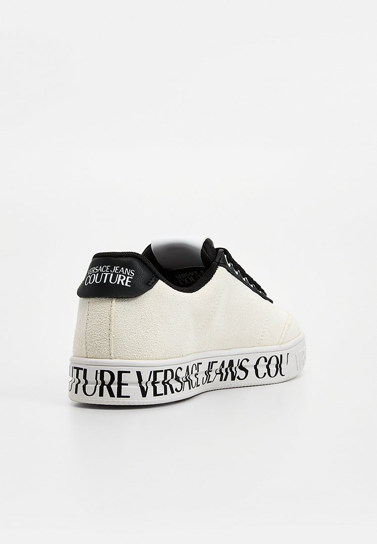 Zapatilla deportiva de ante blanca con un diseño de cordones negros, suela blanca y el texto en negrita negro "VERSACE JEANS COUTURE" en el lateral.
