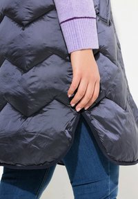 Marineblå pufferväst med chevronmönster, med rundad nederkant. En lavendelfärgad ribbad tröja syns delvis ovanför mörka jeans.