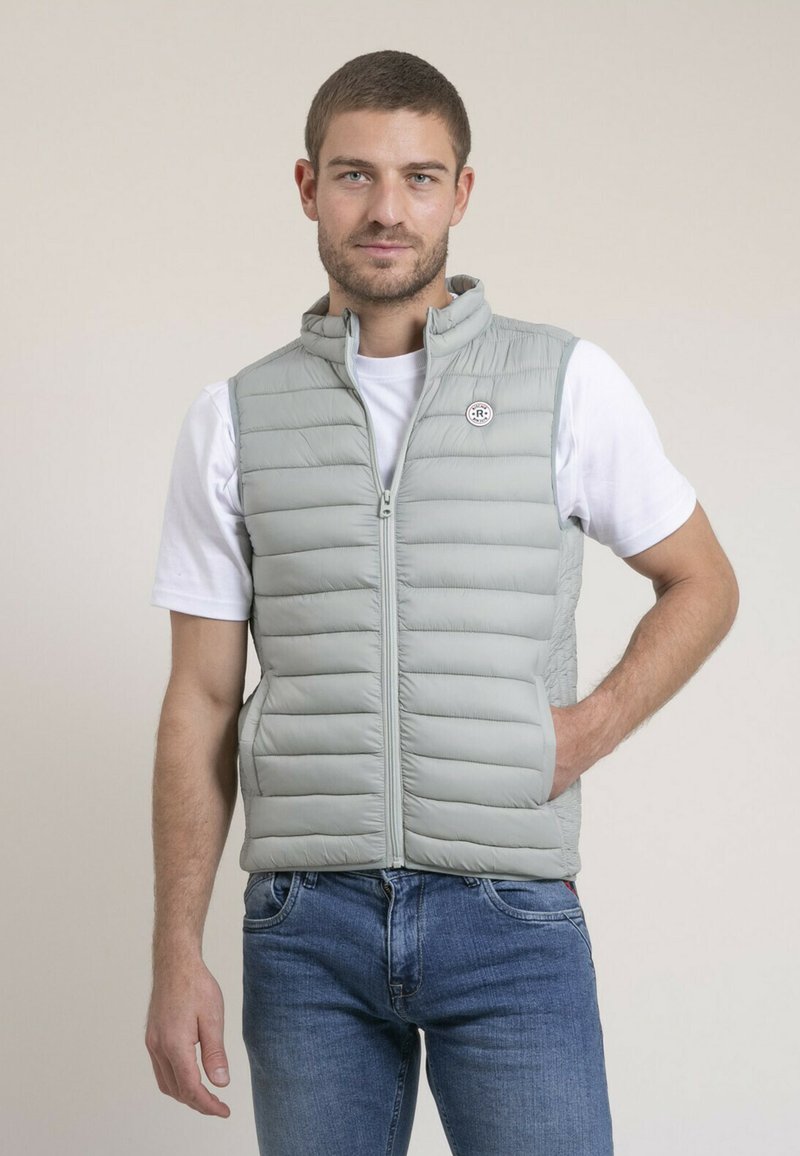 Gilet matelassé gris clair avec un quilting horizontal, col montant, fermeture éclair à l'avant et patch logo. Porté par-dessus un t-shirt blanc et un jean bleu.