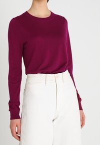 Magenta långärmad tröja med rund halsringning, tillverkad av mjukt stickat tyg. Matchas med vit högmidjad vida byxor.