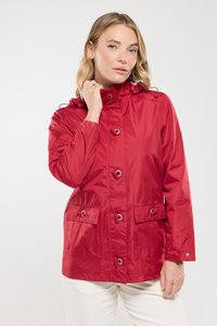 Femme blonde ajuste le col d'une veste rouge à boutons avec des poches avant, portant un pantalon blanc sur fond clair.