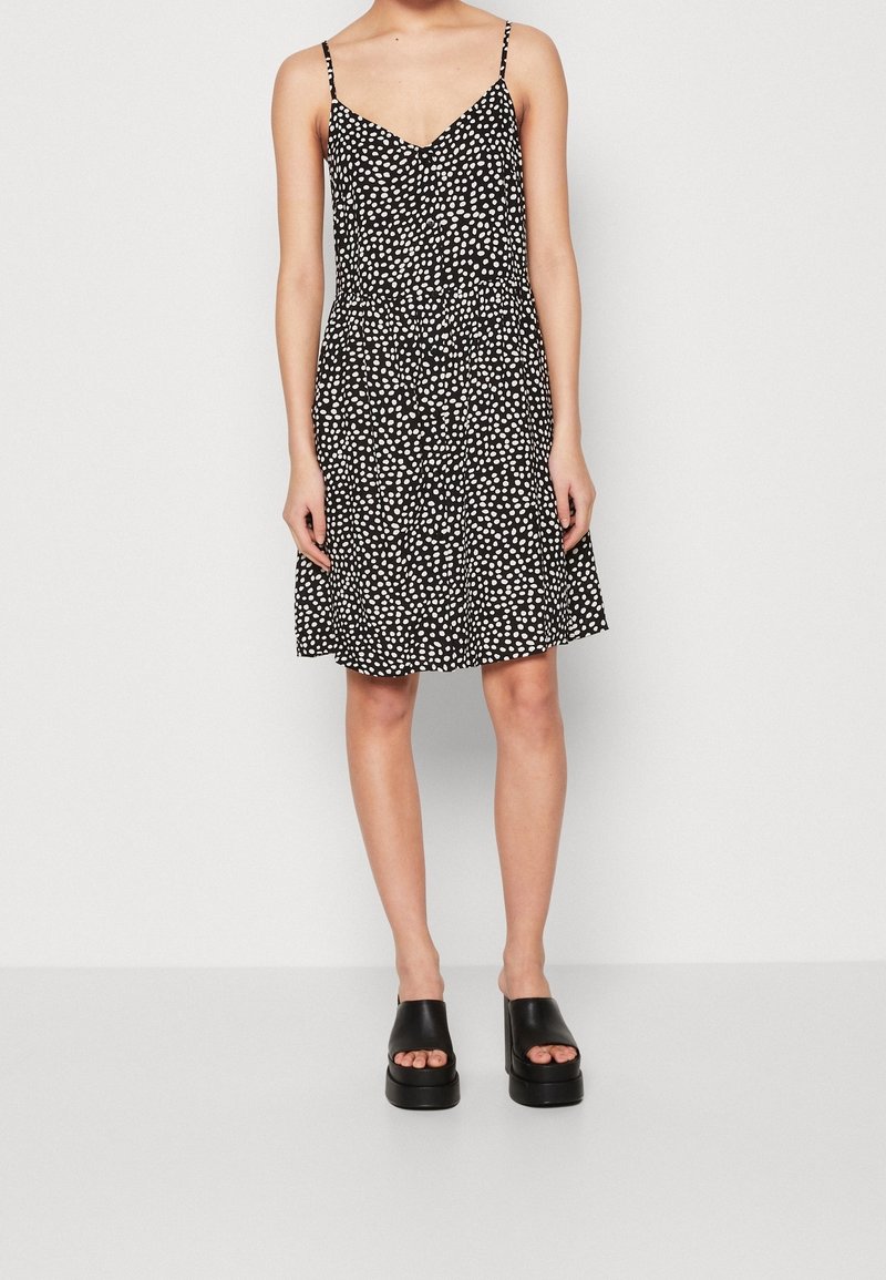 Femme portant une robe noire sans manches à pois blancs et des sandales noires à plateforme à bouts ouverts, debout devant un fond uni.