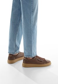 Sneakers in pelle marrone con suola in gomma beige, caratterizzati da un design con lacci e una texture liscia, abbinati a jeans in denim azzurro chiaro.