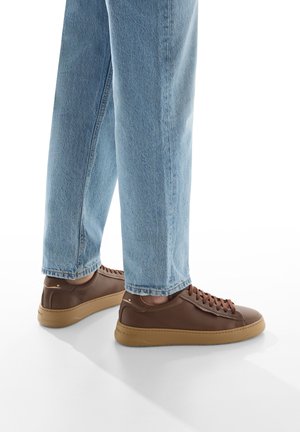 Sneakers in pelle marrone con suola in gomma beige, caratterizzati da un design con lacci e una texture liscia, abbinati a jeans in denim azzurro chiaro.
