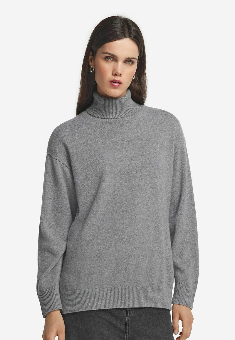 Frau mit langen dunklen Haaren, die silberne Creolen und einen grauen gerippten Rollkragenpullover trägt, vor einem schlichten weißen Hintergrund.