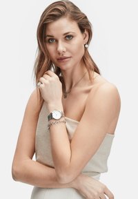 Montre en argent avec un cadran rond blanc et un bracelet à maillons en métal, portée avec un bracelet en chaîne en argent et des colliers superposés.