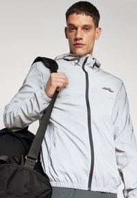 Leichte, graue Zip-Jacke mit Kapuze, elastischen Bündchen und einem schwarzen Reißverschluss. Verfügt über ein Logo auf der Brust. Das Modell hält eine schwarze Tasche.