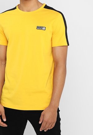 Man wearing a yellow Puma T-shirt with black stripes on shoulders and black pants, standing with arms relaxed.