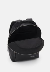 Zaino nero realizzato in tessuto con chiusura a zip. Presenta un ampio interno, una parte superiore arrotondata e un logo in rilievo sulla parte anteriore.
