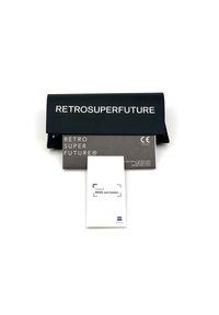 Čierny puzdro na okuliare s textom "RETROSUPERFUTURE" na prednej strane, v sprievode šedej karty a bieleho brožúry označenej "Originálne slnečné šošovky ZEISS."