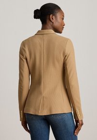 Bēša žakete ar pieguļošu dizainu, teksturētu audumu, ar iegrieztu apkakli un zelta pogu akcentiem piedurknēs. Valkāta kopā ar zilajām džinsām.