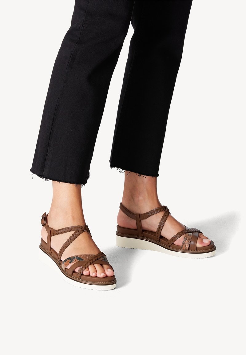 Personne portant un pantalon court noir et des sandales marron à brides tressées avec semelles blanches sur fond blanc.
