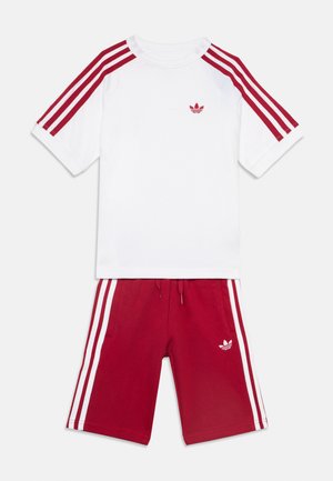 Zestaw dziecięcy Adidas z białą koszulką z czerwonymi paskami na rękawach i dopasowanymi czerwonymi spodenkami z białymi paskami po bokach.